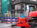 A construction site with a red construction vehicle in the foreground, surrounded by safety barriers and fencing. The building in the background has scaffolding and green netting, indicating ongoing work. There are advertisements visible through the building windows and a bicycle parked nearby.