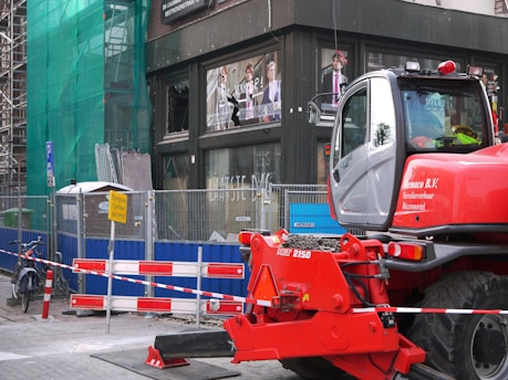 A construction site with a red construction vehicle in the foreground, surrounded by safety barriers and fencing. The building in the background has scaffolding and green netting, indicating ongoing work. There are advertisements visible through the building windows and a bicycle parked nearby.