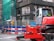 A construction site with a red construction vehicle in the foreground, surrounded by safety barriers and fencing. The building in the background has scaffolding and green netting, indicating ongoing work. There are advertisements visible through the building windows and a bicycle parked nearby.