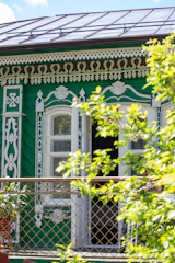 A decorative green wooden house with intricate white and brown carvings above the windows. There are open windows and a lattice fence in front. Lush green leaves partially obscure the view, adding a natural element.