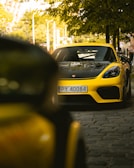 A yellow sports car is parked on a cobblestone street under a tree. The car's license plate reads 'PY 40084'. The environment is urban with buildings and streetlights in the background. The scene has a sunny ambiance, with sunlight filtering through the leaves.