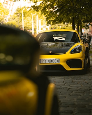 A yellow sports car is parked on a cobblestone street under a tree. The car's license plate reads 'PY 40084'. The environment is urban with buildings and streetlights in the background. The scene has a sunny ambiance, with sunlight filtering through the leaves.