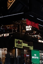 The image features a lively street scene with illuminated signs for several businesses, including 'Kingfisher', 'Just BLR', 'Wow Momo', and 'Mufti'. The signs are vibrant and colorful, with decorations like string lights visible. The storefronts suggest a bustling urban environment at night.