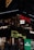 The image features a lively street scene with illuminated signs for several businesses, including 'Kingfisher', 'Just BLR', 'Wow Momo', and 'Mufti'. The signs are vibrant and colorful, with decorations like string lights visible. The storefronts suggest a bustling urban environment at night.