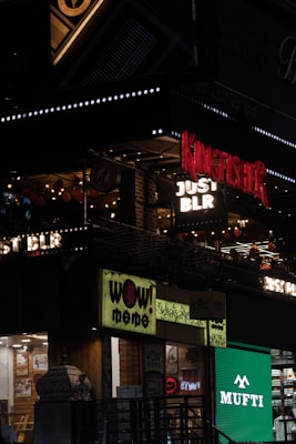 The image features a lively street scene with illuminated signs for several businesses, including 'Kingfisher', 'Just BLR', 'Wow Momo', and 'Mufti'. The signs are vibrant and colorful, with decorations like string lights visible. The storefronts suggest a bustling urban environment at night.