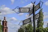 A picturesque scene featuring an ornate street signpost against a backdrop of a historical building with a tall, red brick tower. The signpost, crafted with decorative ironwork, has multiple directional signs in Russian, pointing in various directions. Lush green trees and a partly cloudy blue sky add to the tranquil setting.
