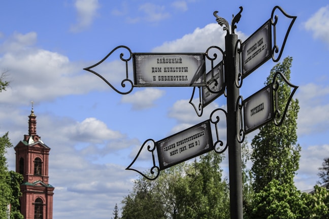 A picturesque scene featuring an ornate street signpost against a backdrop of a historical building with a tall, red brick tower. The signpost, crafted with decorative ironwork, has multiple directional signs in Russian, pointing in various directions. Lush green trees and a partly cloudy blue sky add to the tranquil setting.