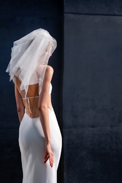 A person wearing a white wedding gown with an open back design and a fine veil over their head stands in front of a dark, textured wall. Their posture is slightly turned away, with one arm casually resting at their side, showing red nail polish.