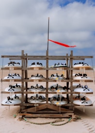 A wooden rack on a sandy beach displays multiple rows of white sandboards, each with black straps. A red flag flutters on a pole above the rack against a cloudy sky.