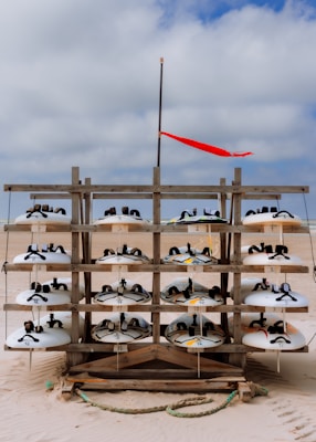 A wooden rack on a sandy beach displays multiple rows of white sandboards, each with black straps. A red flag flutters on a pole above the rack against a cloudy sky.