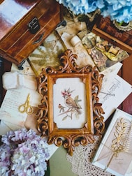 An ornate wooden frame containing a bird embroidery is surrounded by vintage items including old letters, sheet music, a small pair of gold scissors, and a bouquet of hydrangeas. The scene is set on a wooden surface, with additional items like a magnifying glass and a wooden chest adding to the antique feel.