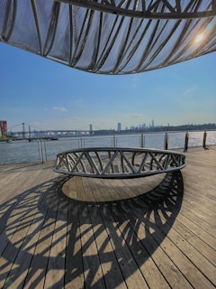 An outdoor wooden deck area with modern metal circular seating and an overhead architectural structure creating intriguing shadows. In the background, a body of water is visible with a bridge spanning across it. A city skyline with tall buildings is seen in the distance under a clear sky.