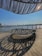 An outdoor wooden deck area with modern metal circular seating and an overhead architectural structure creating intriguing shadows. In the background, a body of water is visible with a bridge spanning across it. A city skyline with tall buildings is seen in the distance under a clear sky.