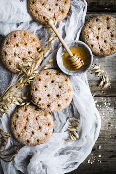 Round, rustic crackers are artfully arranged on a wooden surface, accompanied by a small bowl of honey with a wooden honey dipper. Delicate sprigs of oats and a thin, translucent fabric add a natural and textured element to the composition.