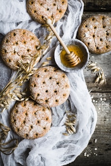Round, rustic crackers are artfully arranged on a wooden surface, accompanied by a small bowl of honey with a wooden honey dipper. Delicate sprigs of oats and a thin, translucent fabric add a natural and textured element to the composition.