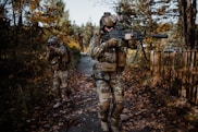 Two individuals in camouflage gear walk through a wooded area, carrying rifles. The surroundings are filled with autumn foliage, and the path beneath them is covered in fallen leaves. One person is aiming, while the other appears to be scanning the environment.