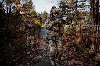 Two individuals in camouflage gear walk through a wooded area, carrying rifles. The surroundings are filled with autumn foliage, and the path beneath them is covered in fallen leaves. One person is aiming, while the other appears to be scanning the environment.