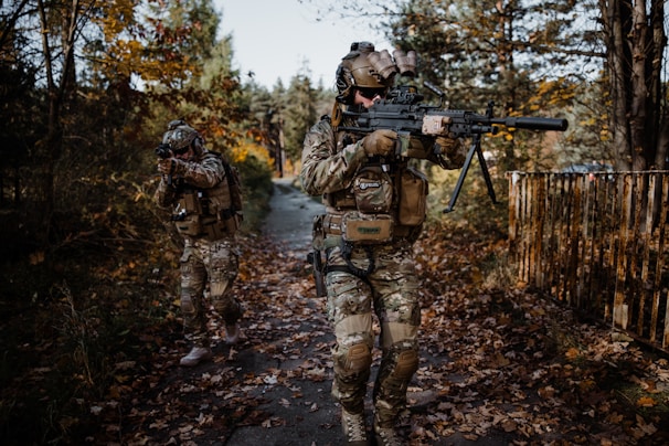 Two individuals in camouflage gear walk through a wooded area, carrying rifles. The surroundings are filled with autumn foliage, and the path beneath them is covered in fallen leaves. One person is aiming, while the other appears to be scanning the environment.