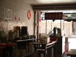 A small street food stand with a variety of kitchen supplies, utensils, and containers arranged on metal tables. There are red signs with Chinese characters hanging on the walls and on a cart. The setup has an informal, makeshift appearance, typical of a local eatery or snack bar.