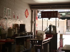 A small street food stand with a variety of kitchen supplies, utensils, and containers arranged on metal tables. There are red signs with Chinese characters hanging on the walls and on a cart. The setup has an informal, makeshift appearance, typical of a local eatery or snack bar.