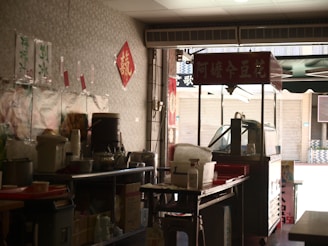A small street food stand with a variety of kitchen supplies, utensils, and containers arranged on metal tables. There are red signs with Chinese characters hanging on the walls and on a cart. The setup has an informal, makeshift appearance, typical of a local eatery or snack bar.