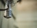 A metallic combination padlock hangs from a looped chain, with the numbers 3, 4, 5, 6 displayed on its dials. The background is blurred, giving focus to the lock which appears slightly worn.