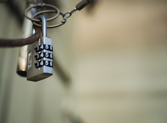 A metallic combination padlock hangs from a looped chain, with the numbers 3, 4, 5, 6 displayed on its dials. The background is blurred, giving focus to the lock which appears slightly worn.