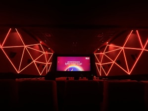An interior view of a modern movie theater featuring geometric red neon lights on the side walls. The room is dark with rows of seats visible in the foreground. The screen displays a message that says 'Welcome to the movies, let the magic begin!'