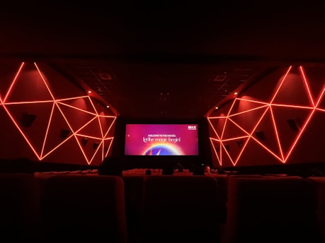 An interior view of a modern movie theater featuring geometric red neon lights on the side walls. The room is dark with rows of seats visible in the foreground. The screen displays a message that says 'Welcome to the movies, let the magic begin!'