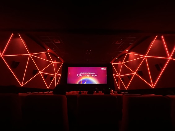 An interior view of a modern movie theater featuring geometric red neon lights on the side walls. The room is dark with rows of seats visible in the foreground. The screen displays a message that says 'Welcome to the movies, let the magic begin!'