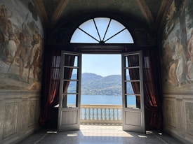 A room with classical wall murals on both sides leads to an open set of wooden doors. Beyond the doors, a stunning view of a tranquil lake and distant hills is visible. The ceiling features ornate designs, and the natural light from outside enhances the rich colors in the room.