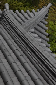 A detailed view of traditional tiled roofing with overlapping grey tiles, highlighted by the shadows and textures of the tile design. The roof is set against a backdrop of lush green foliage, providing a natural contrast.