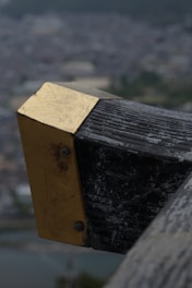 A close-up view of a weathered metal and wooden structure with a brass or gold-colored plate attached to it using screws. The background features a blurred cityscape or landscape, suggesting the structure is elevated above ground level.