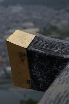 A close-up view of a weathered metal and wooden structure with a brass or gold-colored plate attached to it using screws. The background features a blurred cityscape or landscape, suggesting the structure is elevated above ground level.