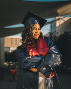 A person wearing a graduation cap and gown, with a confident pose, is standing in an outdoor setting. The gown is dark blue with a red sash. Sunlight casts dramatic shadows around the figure, and buildings are visible in the background.