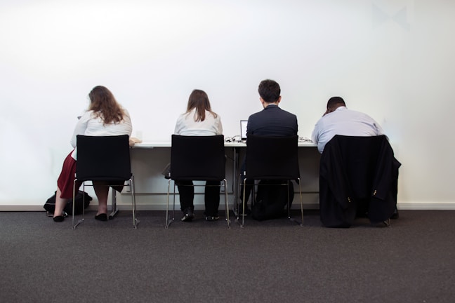Four people sit at a table, seen from behind, in a plain room. They are dressed in business attire and appear to be focused on work at a desk with laptops and papers.
