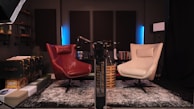 A cozy and modern recording studio setup featuring two swivel chairs, one red and one cream, placed on a patterned rug. The room includes acoustic panels on the walls, shelves with decor items, books, a microphone arm, and soft ambient lighting. A vintage-style suitcase and several boxes are stacked on the left.