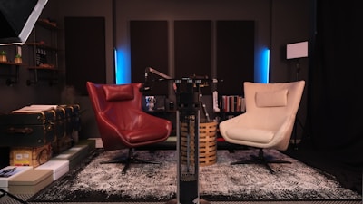 A cozy and modern recording studio setup featuring two swivel chairs, one red and one cream, placed on a patterned rug. The room includes acoustic panels on the walls, shelves with decor items, books, a microphone arm, and soft ambient lighting. A vintage-style suitcase and several boxes are stacked on the left.
