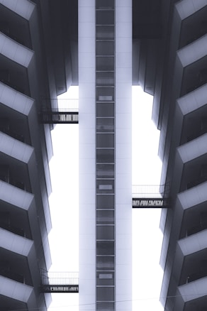 A symmetrical view of a modern high-rise building with a central elevator shaft. The structure features sharp geometric lines, contrasting with the sky visible through narrow gaps. Balconies with metal railings are present on both sides, enhancing the sense of depth.
