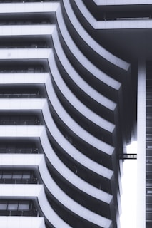 A modern architectural structure features a series of sweeping, curved balconies that create a rhythmic pattern. The design incorporates smooth white surfaces contrasted with dark shadows, giving a sense of depth and sophistication. The repeating curves are both functional and artistic, suggesting a contemporary approach to building design.