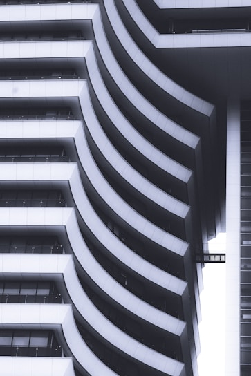 A modern architectural structure features a series of sweeping, curved balconies that create a rhythmic pattern. The design incorporates smooth white surfaces contrasted with dark shadows, giving a sense of depth and sophistication. The repeating curves are both functional and artistic, suggesting a contemporary approach to building design.