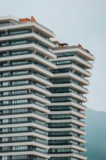 A modern high-rise building with a unique staggered design, featuring large glass windows and horizontal white bands. The structure stands against a backdrop of misty hills, with some construction equipment visible on the rooftop.