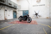 A black motorcycle is parked on a red oriental-style carpet in an industrial-style warehouse. The walls are light-colored with signs of wear, and there's a blue double door in the background. Metal ducts and a sign that reads 'Rauchen Verboten' are visible. A large propeller sculpture is mounted on the wall.