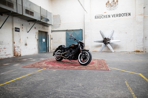 A black motorcycle is parked on a red oriental-style carpet in an industrial-style warehouse. The walls are light-colored with signs of wear, and there's a blue double door in the background. Metal ducts and a sign that reads 'Rauchen Verboten' are visible. A large propeller sculpture is mounted on the wall.