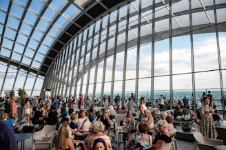 A spacious indoor venue with large, curved glass windows showcasing a panoramic city view. The area is bustling with people engaged in various activities like chatting, sitting, and taking photos. The ceiling has a modern, arched design with metal frames and the space features some plants and minimalistic furniture.