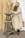 A bearded man wearing a loose white garment is sculpting a clay figure on a tall wooden stool. The scene appears to be set in an artist's studio with a focus on the creative process.