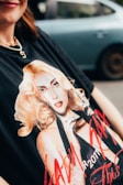 A close-up of a person wearing a black t-shirt featuring a printed graphic of a blonde woman with a striking pose. The text on the shirt includes 'TOUR 2011'. The person is wearing a necklace with a heart pendant, and there is a blurred car in the background.