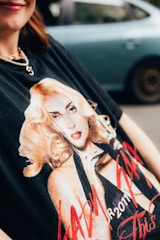 A close-up of a person wearing a black t-shirt featuring a printed graphic of a blonde woman with a striking pose. The text on the shirt includes 'TOUR 2011'. The person is wearing a necklace with a heart pendant, and there is a blurred car in the background.