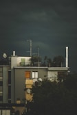 A building with a rooftop garden and several antennas. The structure appears modern, with large windows, and there are trees partially obscuring the lower part of the building. The sky is dark and overcast, suggesting an impending storm.