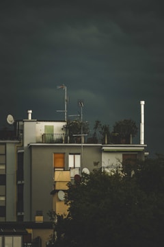 A building with a rooftop garden and several antennas. The structure appears modern, with large windows, and there are trees partially obscuring the lower part of the building. The sky is dark and overcast, suggesting an impending storm.